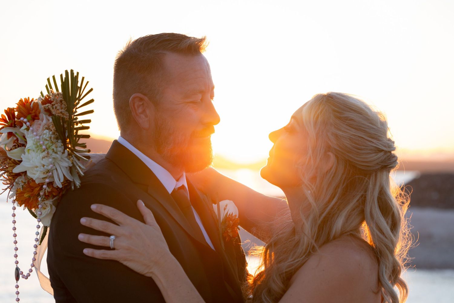 Silhouetted couple embraces and kisses at sunset on their wedding day with ocean backdrop and floral bouquet at the Riviera Lake Havasu