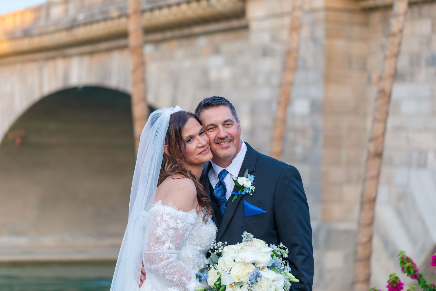 Smiling bride and groom pose together in front of arched bridge at outdoor wedding venue at Shugrues in Lake Havasu by the London Bridge