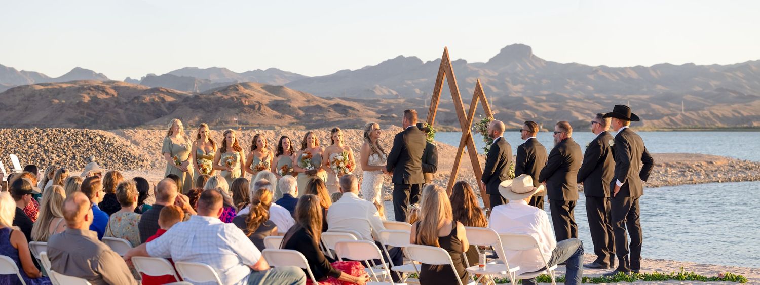Riviera Marina wedding ceremony takes place at sunset by a lake with mountain views as guests watch from white chairs in Lake Havasu