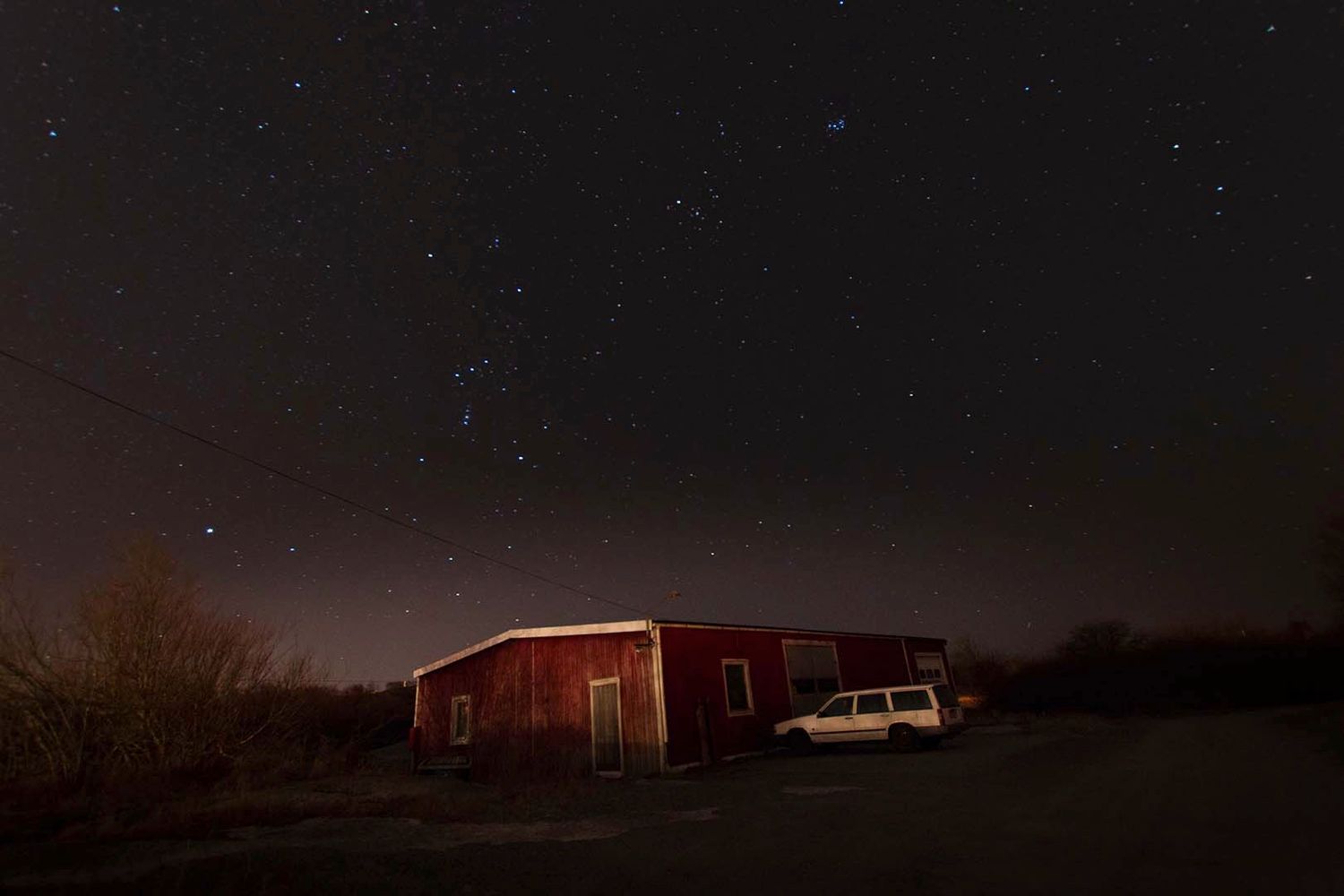 Red barn and parked car under starry night sky in rural setting.
