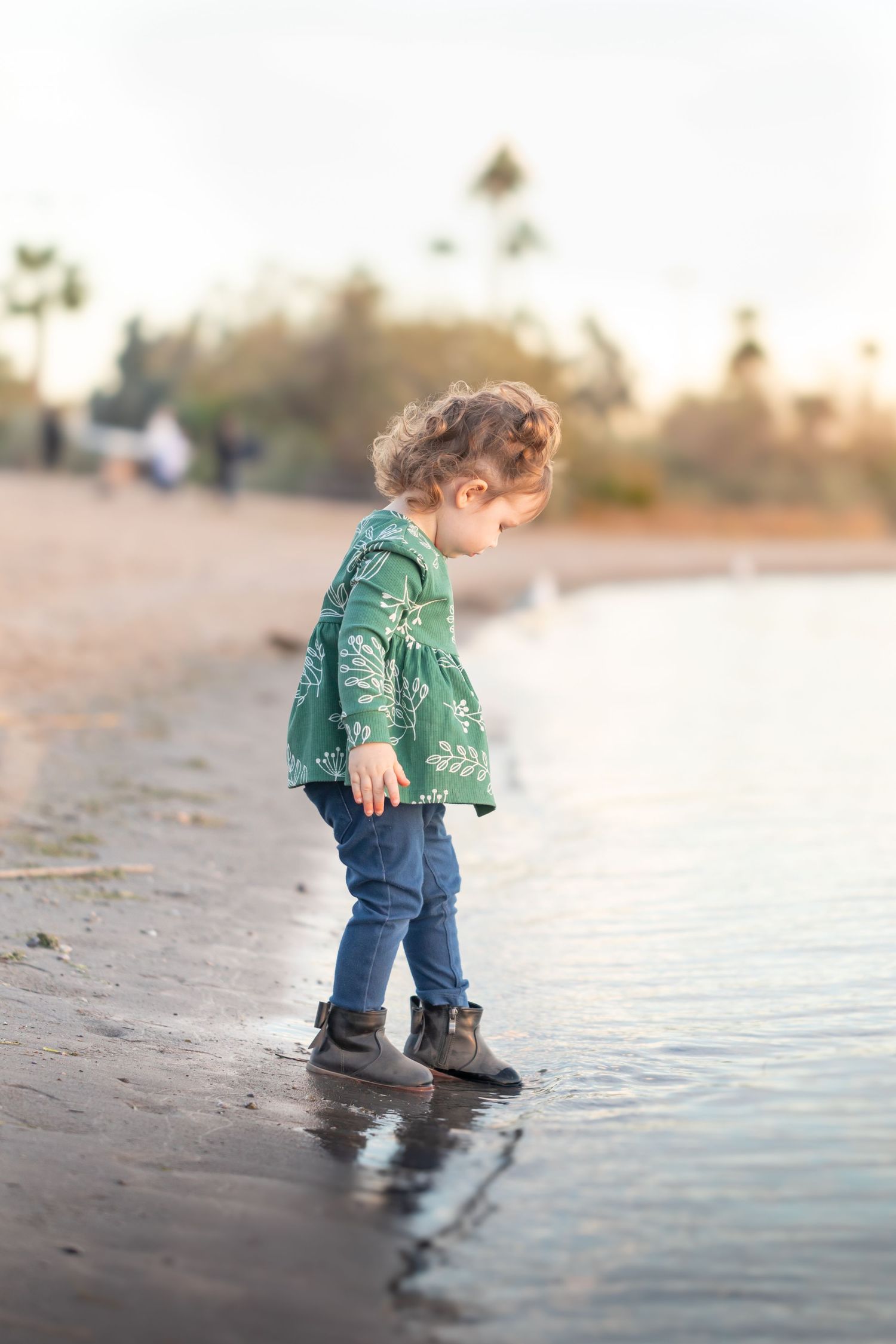 Toddler girl in green floral top and jeans exploring shallow water at a sandy beach during golden hour.