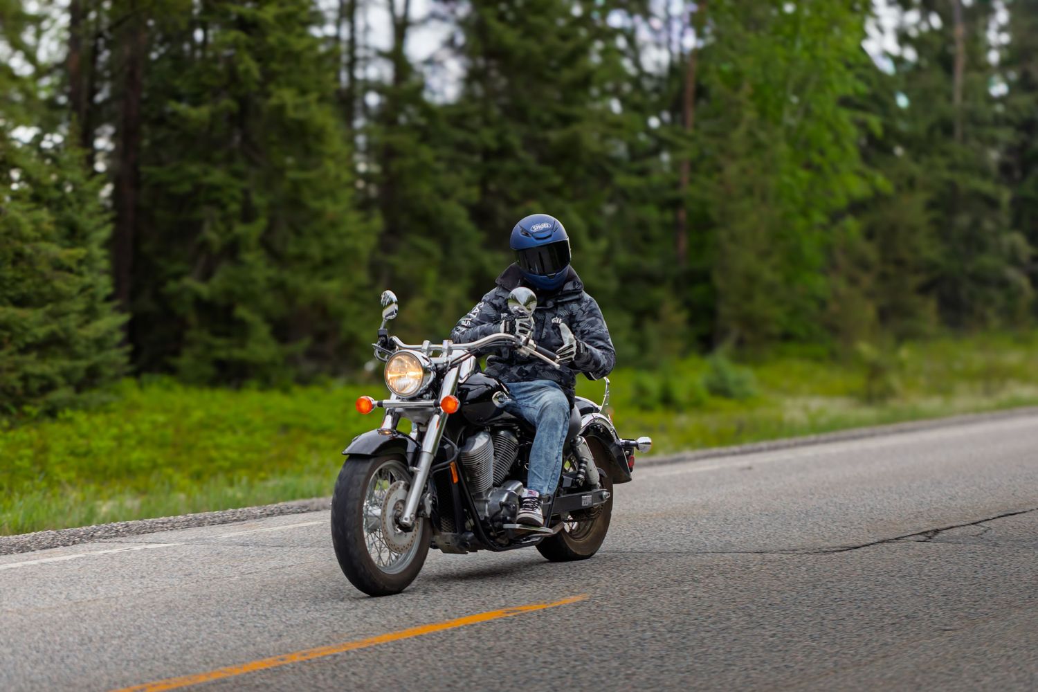 Motorcyclist riding a cruiser motorcycle on a wet road surrounded by lush green forest trees.