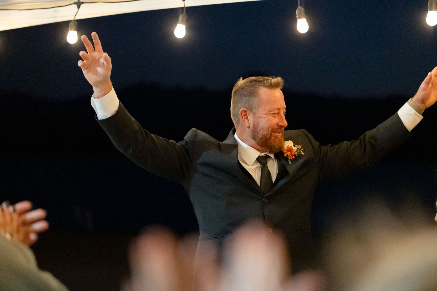 Groomsman in dark suit raises arms triumphantly under string lights at Lake Havasu wedding reception.