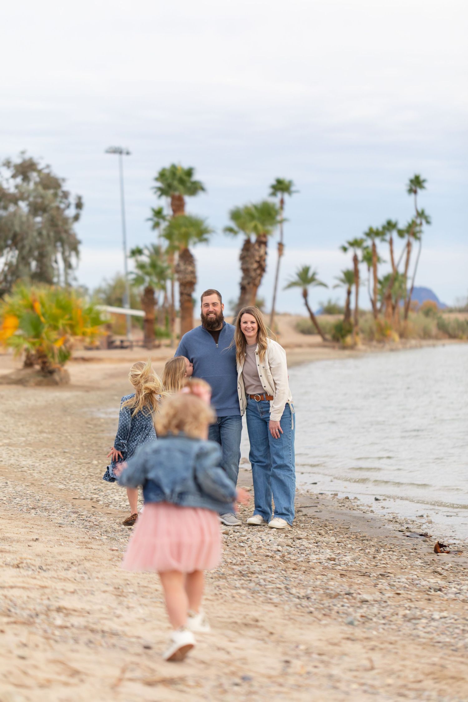 Family of four enjoying a sunny day on a sandy beach with palm trees, two young girls running toward smiling parents.