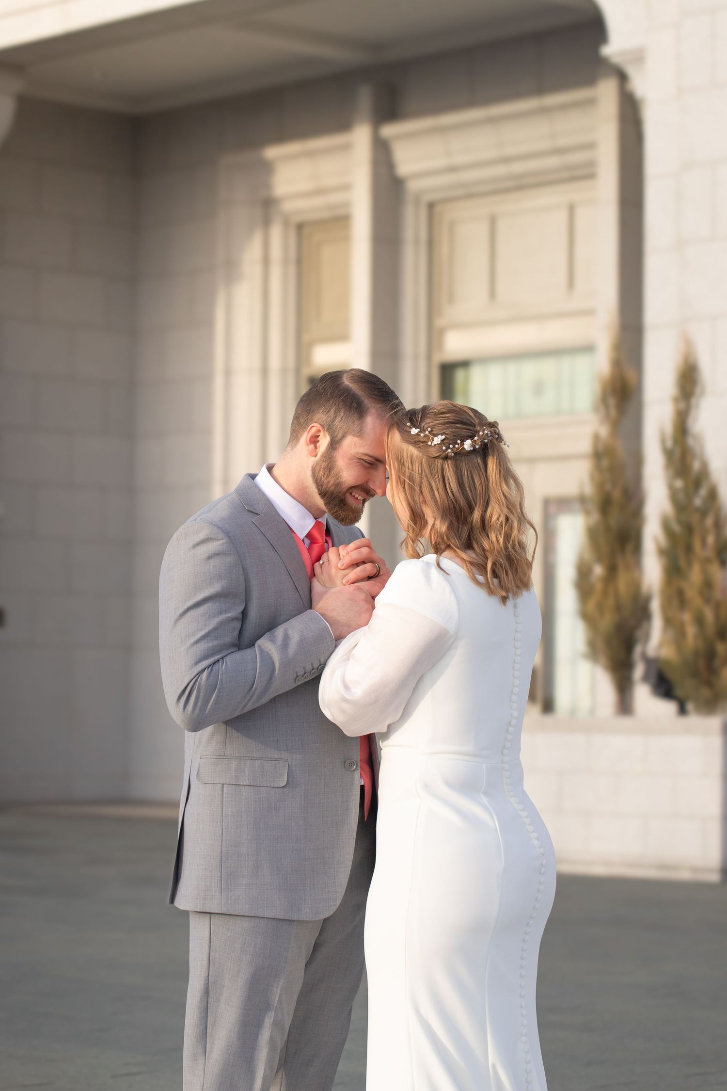Bride and groom in white dress and gray suit with red tie share an intimate moment outdoors near a building.