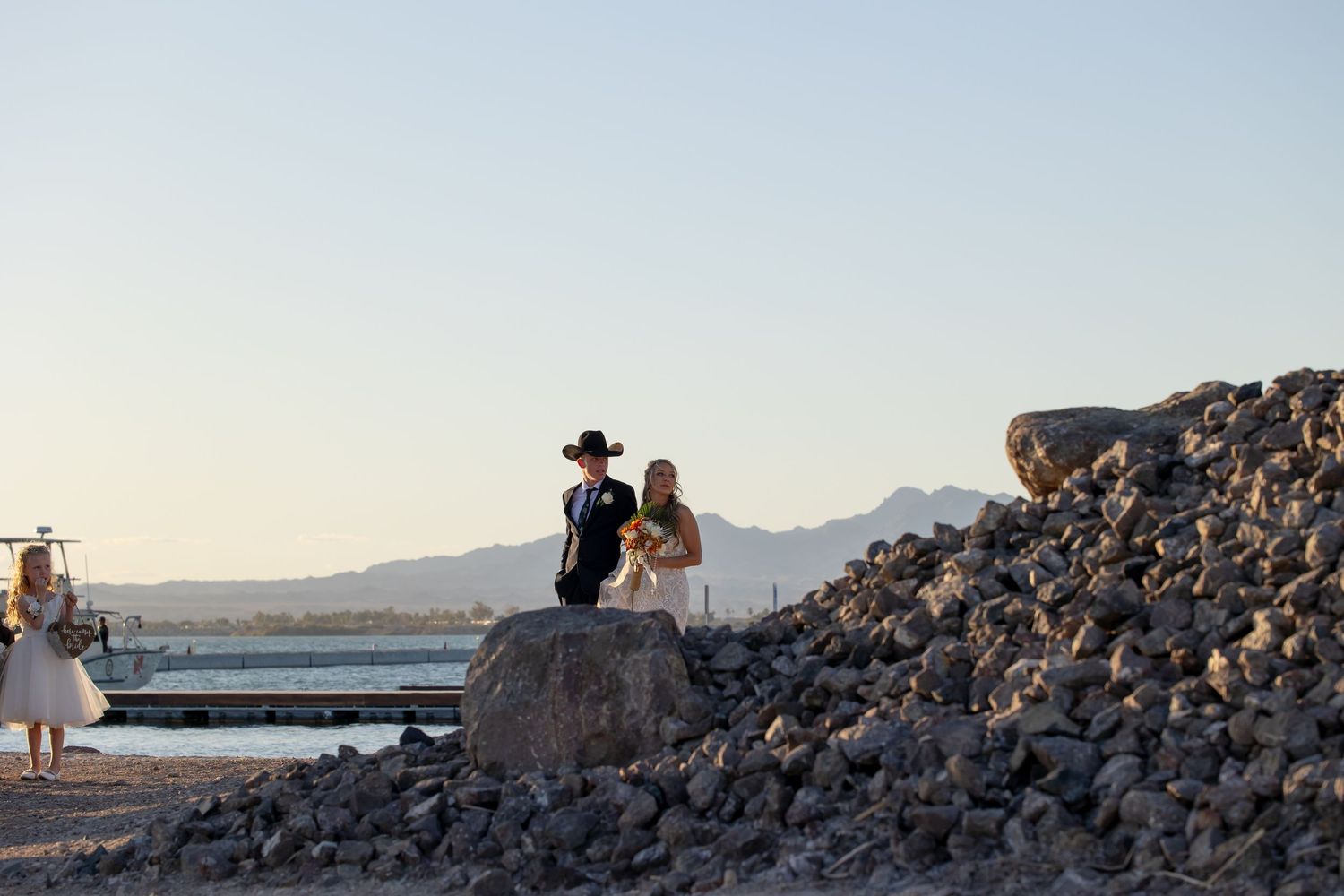 Couple in wedding attire standing on rocky terrain with mountains and clear sky in the background.