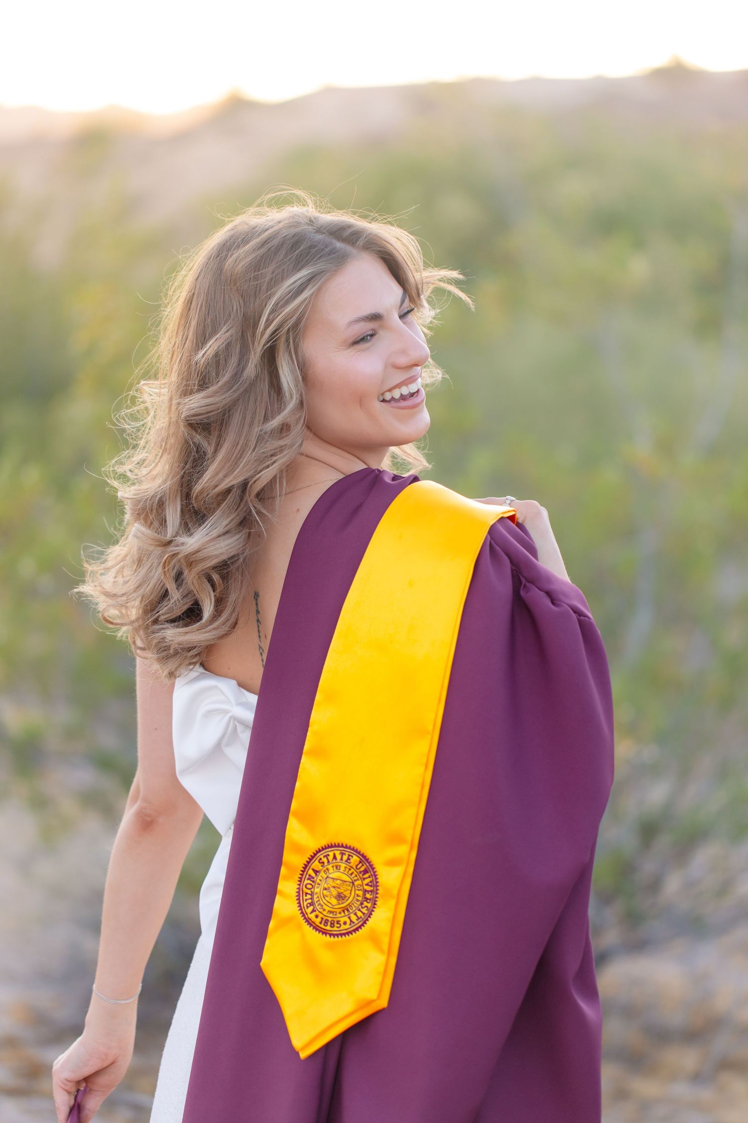 Smiling blonde woman in white dress holding maroon and gold ASU graduation gown and stole outdoors at sunset in Lake Havasu City Arizona
