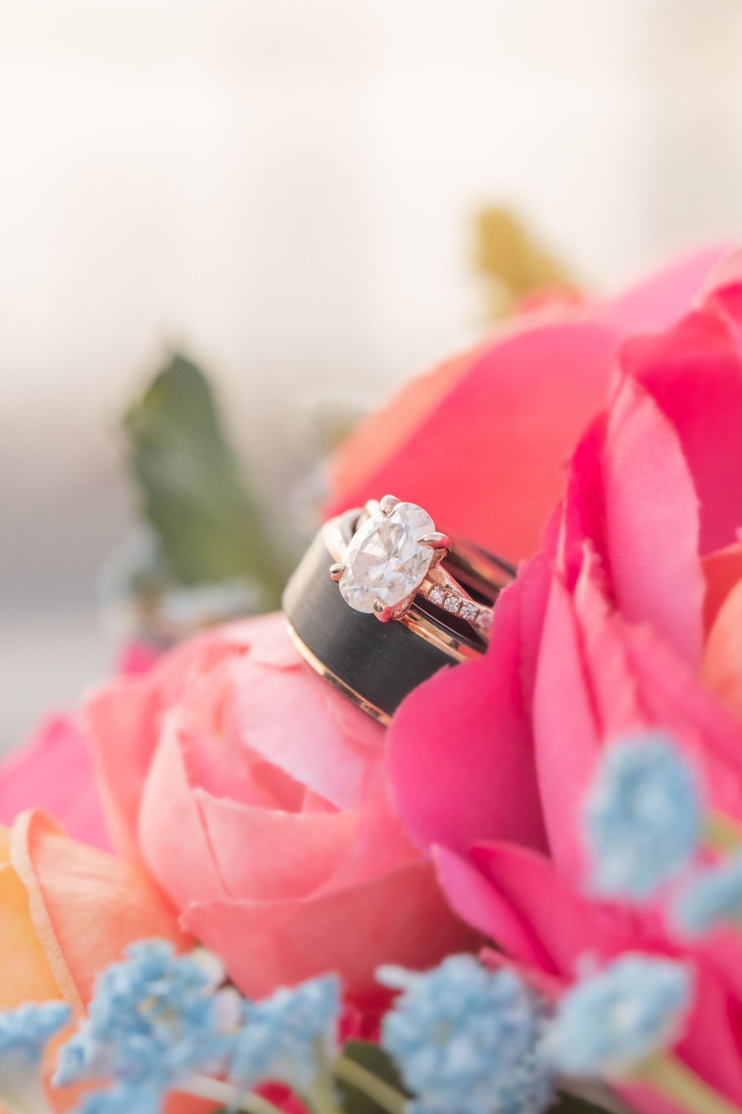Diamond engagement ring and wedding band nestled among pink roses and blue delphinium flowers in a romantic setting.