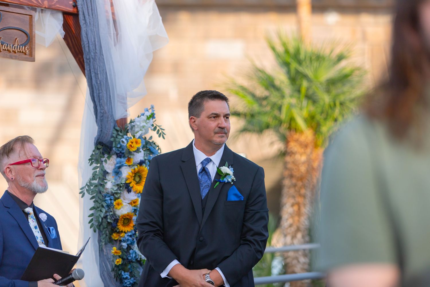 Groom in gray suit with blue tie smiling at wedding ceremony altar decorated with sunflowers outdoors at Shugrues in Lake Havasu