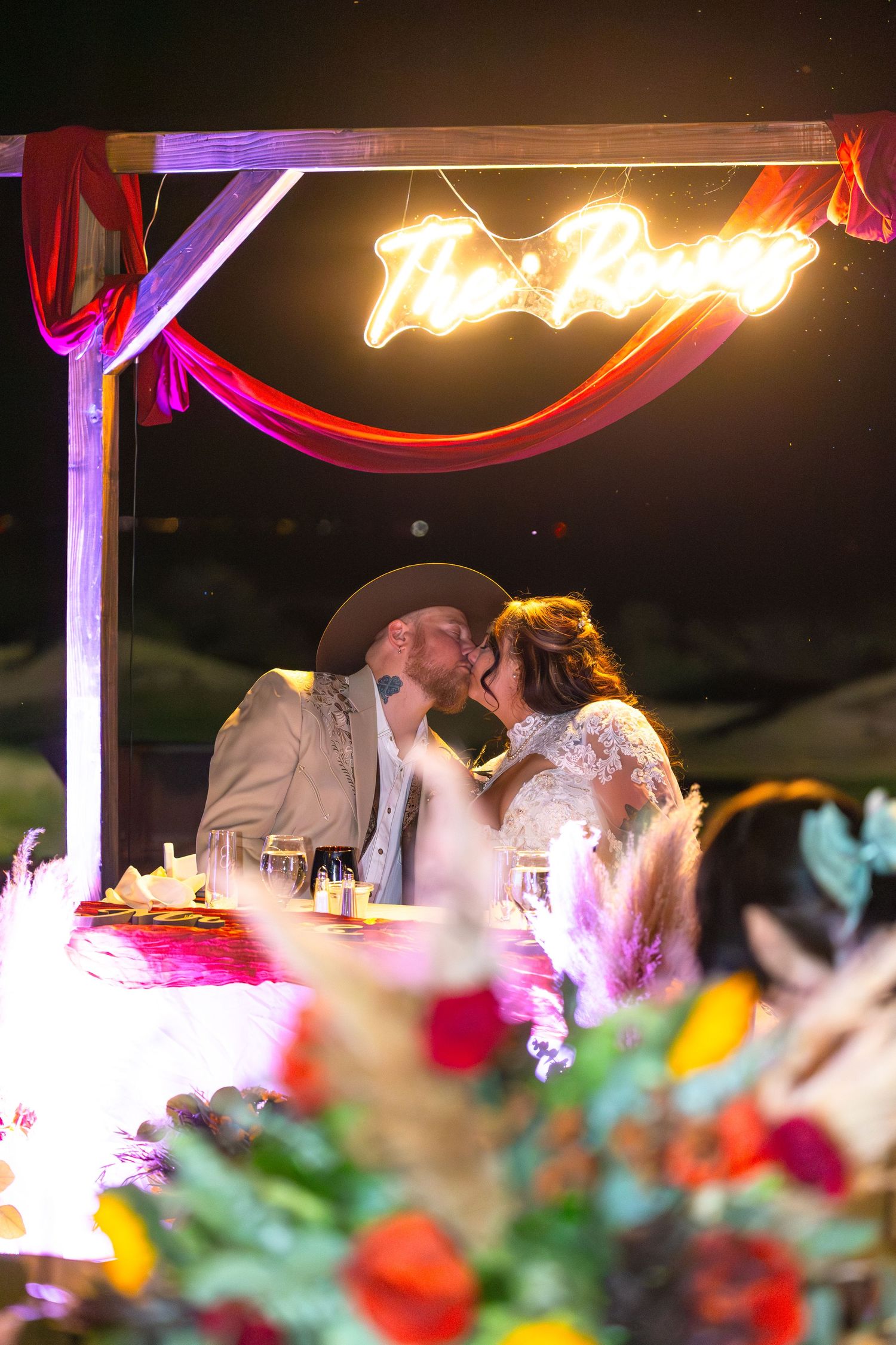 A newlywed couple shares a kiss at their sweetheart table beneath a glowing neon sign at their romantic wedding reception at Iron Wolf