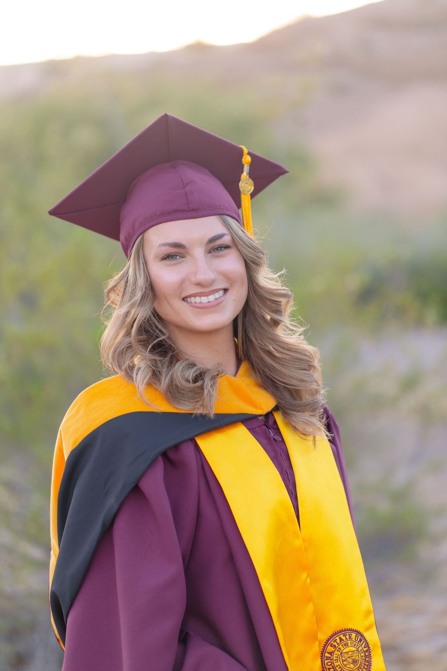 Smiling young woman in maroon and gold graduation cap and gown posing outdoors in natural desert setting.