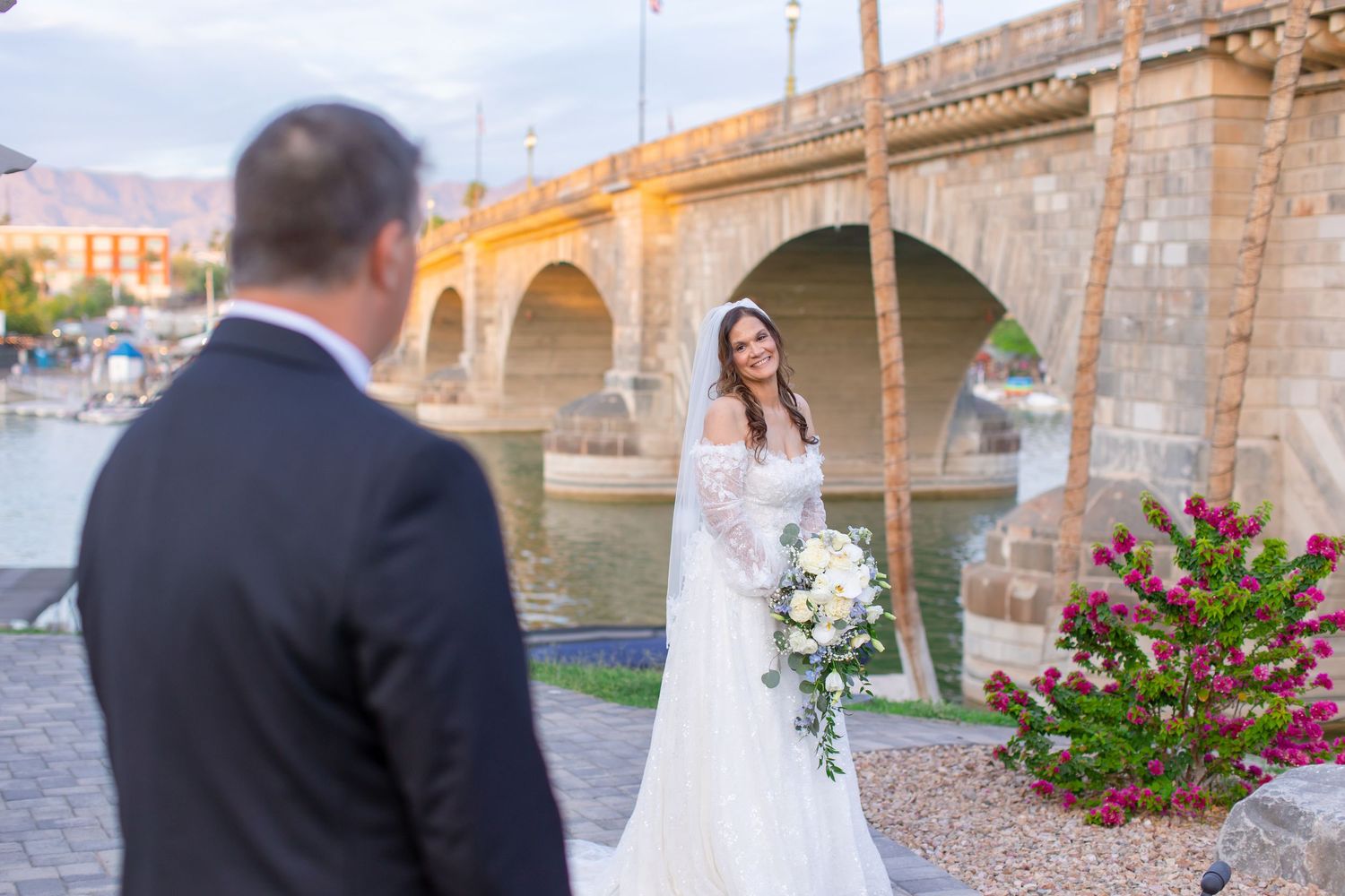 Bride in elegant white lace gown holds bouquet near London Bridge waterfront at sunset during wedding photo session at Shugrues