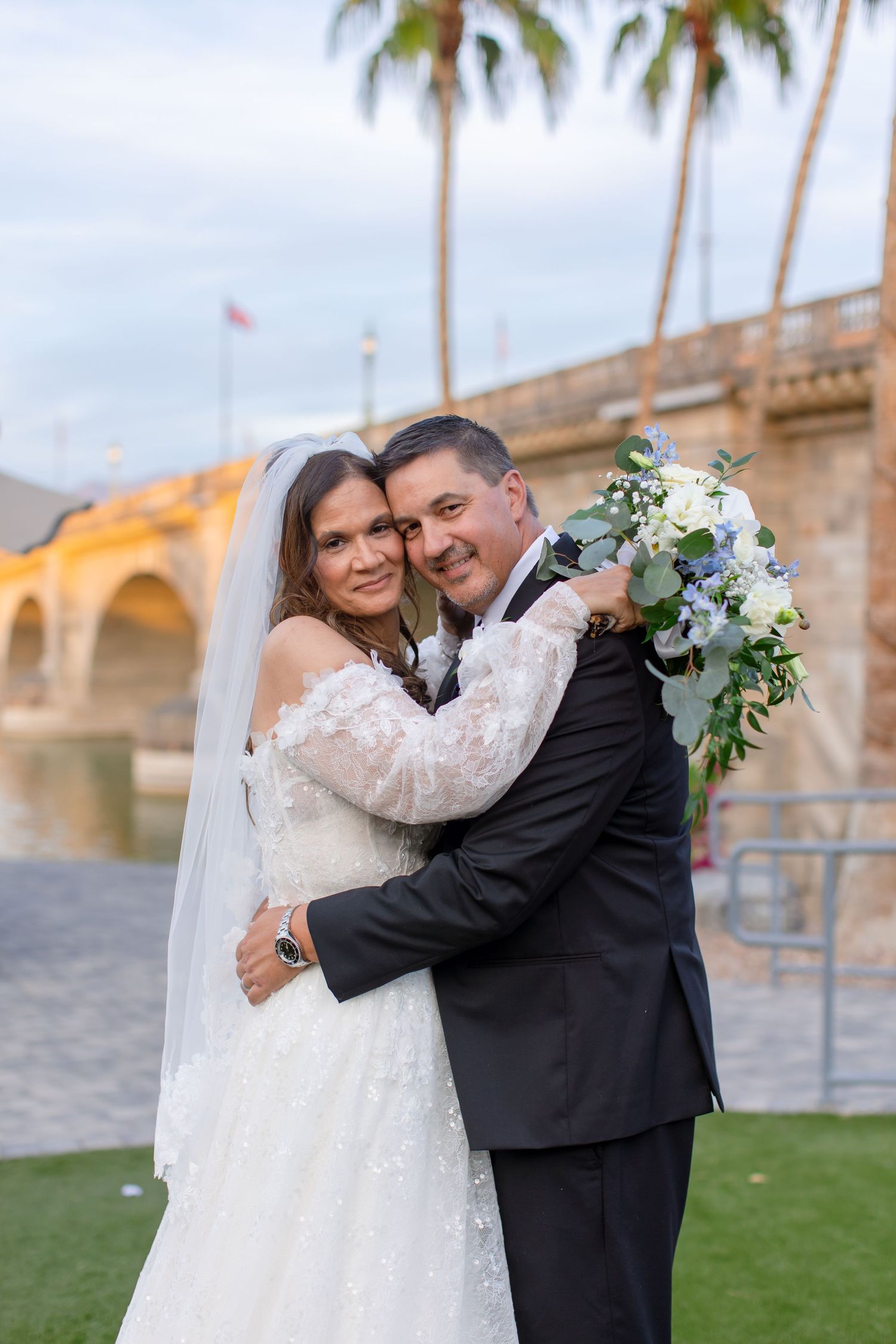 Bride and groom embracing outdoors near the london bridge, holding a white and blue bouquet, with palm trees in the background at Shugrues