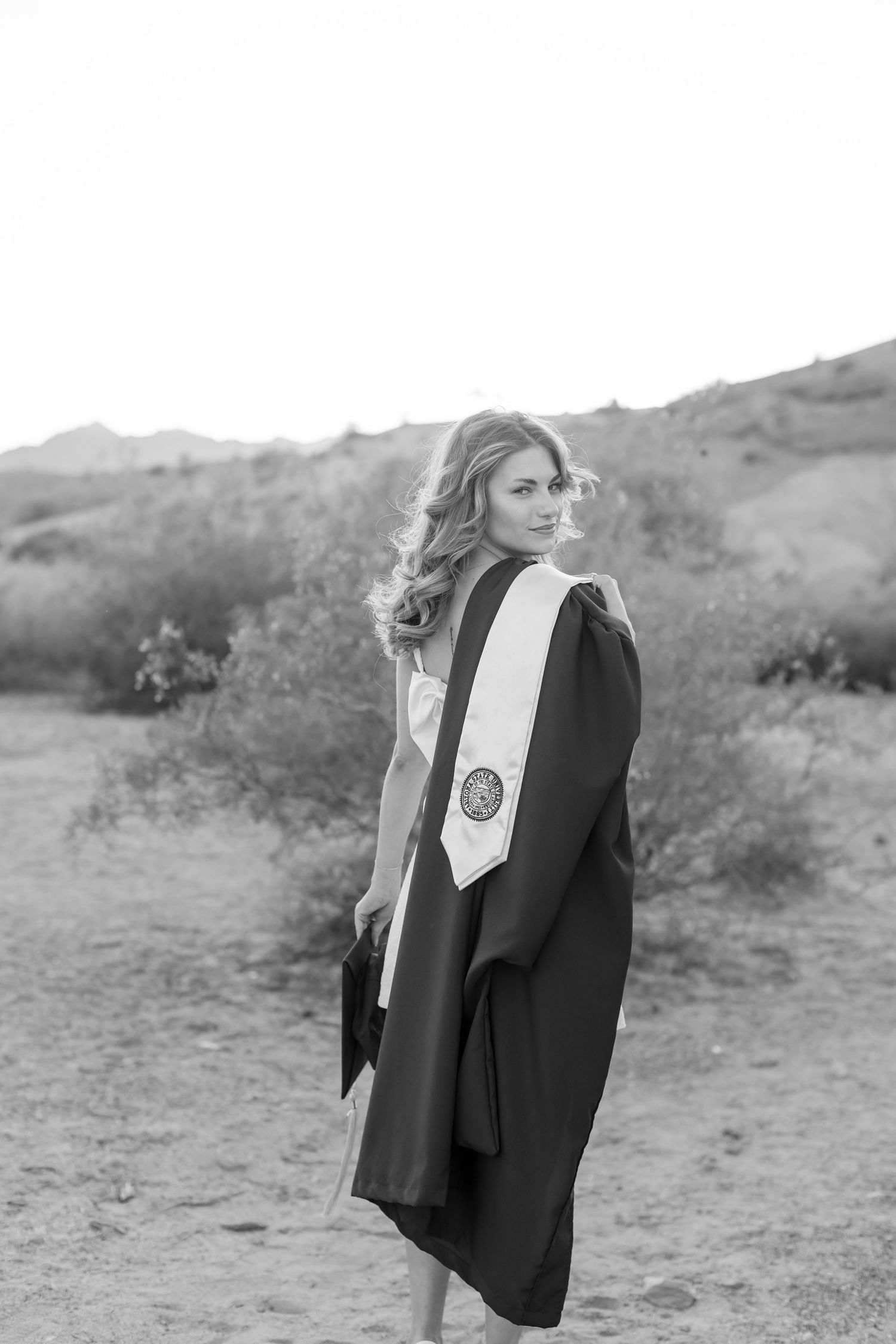 Black and white photo of a young woman in graduation gown and sash posing outdoors in a desert landscape in Lake Havasu City Arizona