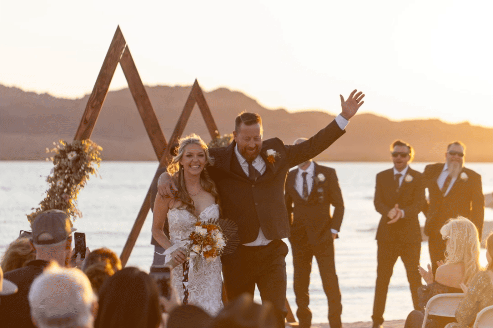 A joyful celebration at a lakeside Riviera Marina wedding ceremony in Lake Havasu City Arizona with a triangular wooden arch at sunset.