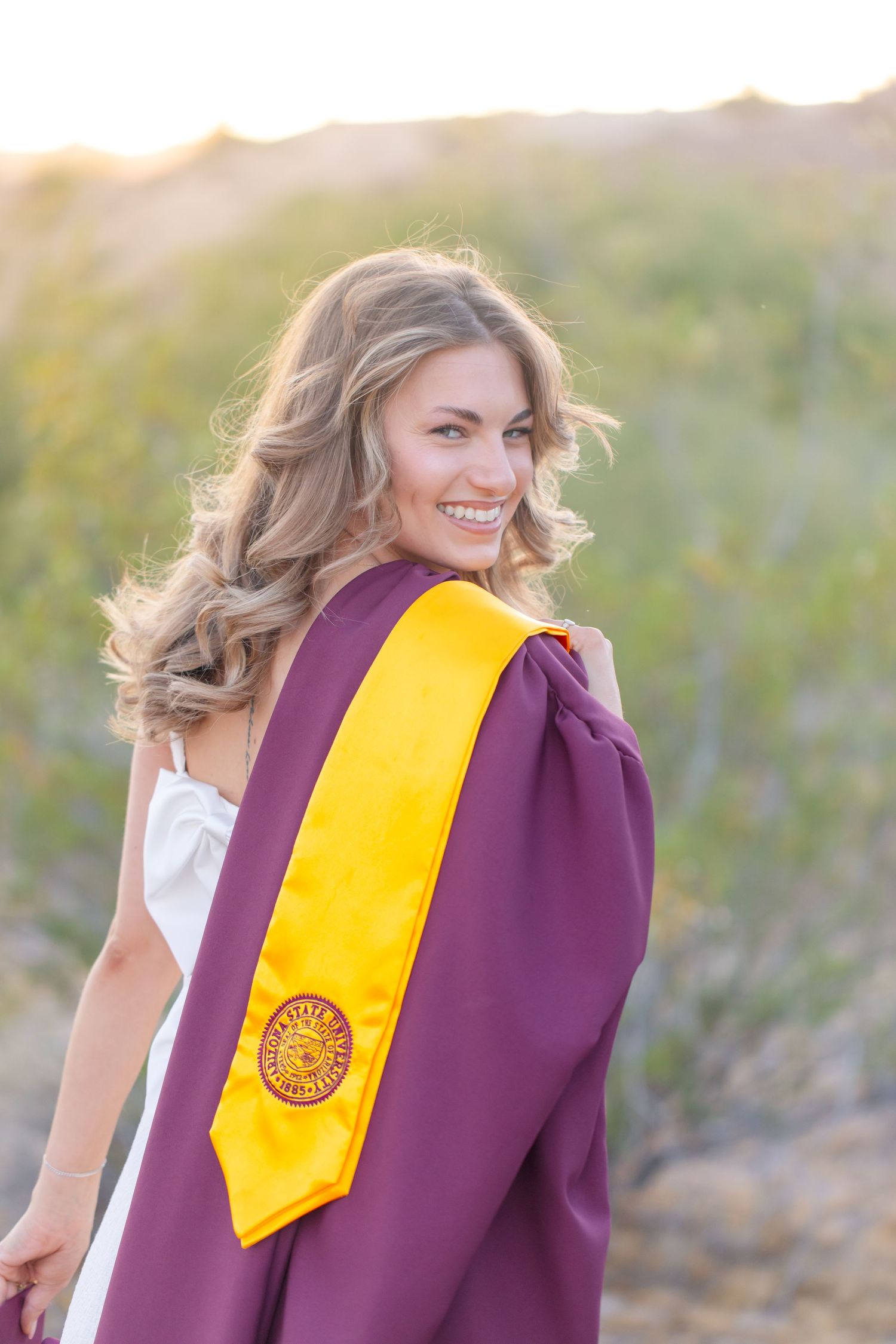 Smiling female graduate in maroon gown and gold ASU sash poses outdoors in golden hour sunlight.