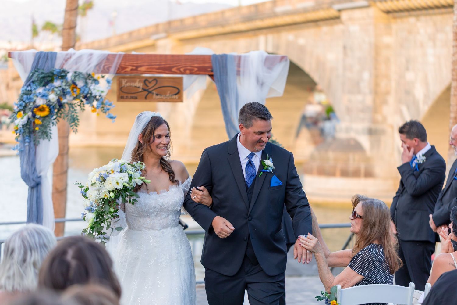 Bride and groom walk down the aisle at an outdoor waterfront wedding ceremony with floral arch at Shugrues in Lake Havasu City Arizona