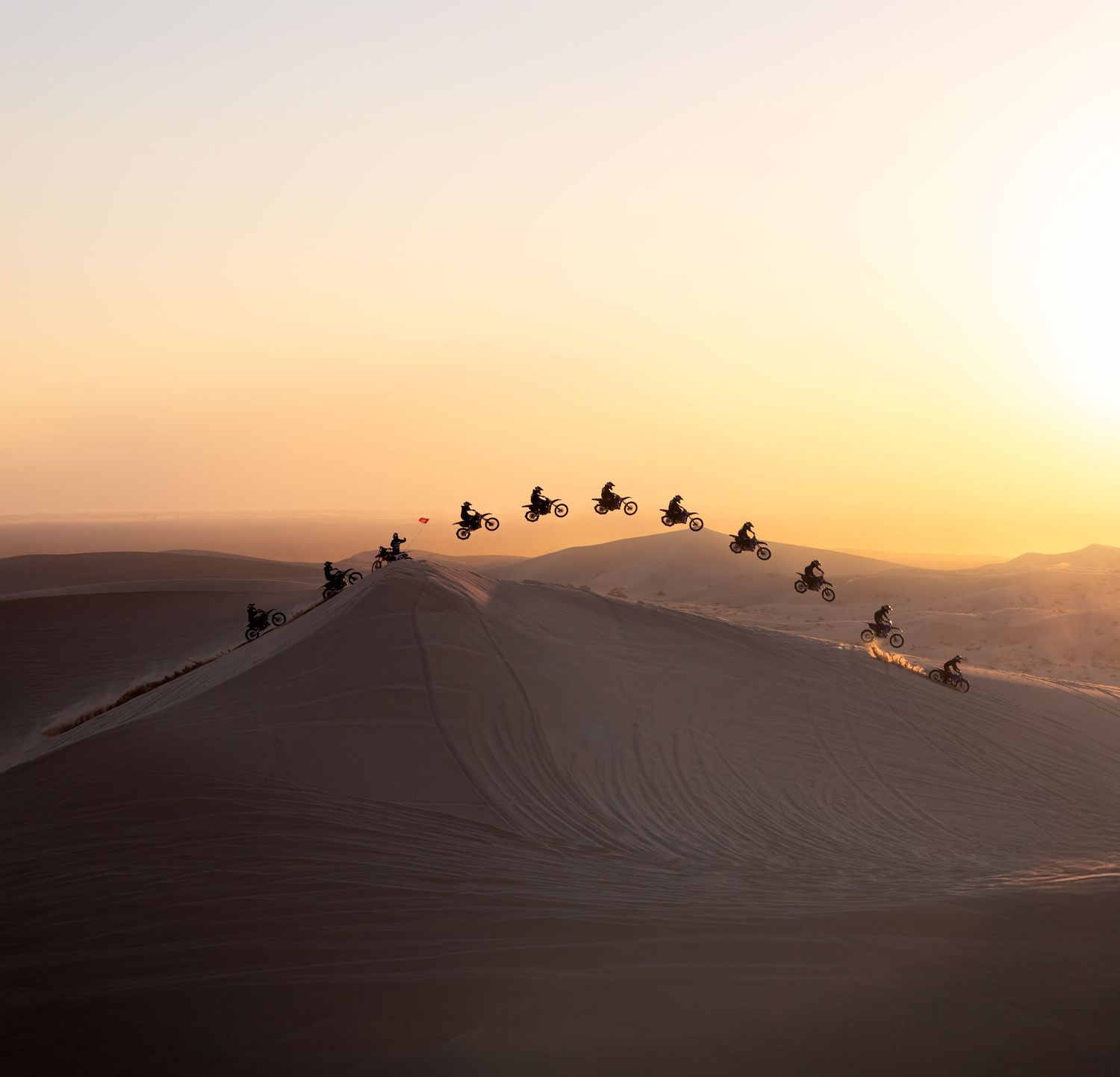 Motorcyclists silhouetted against a golden sunset sky, jumping over a sand dune in the desert.