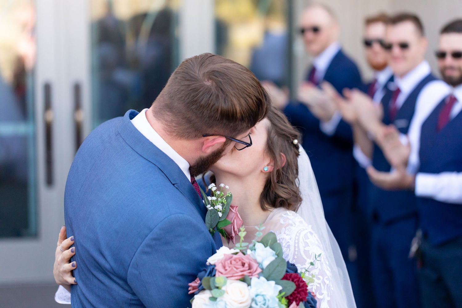Wedding couple sharing romantic kisses with guests and wedding party in formal attire looking on.