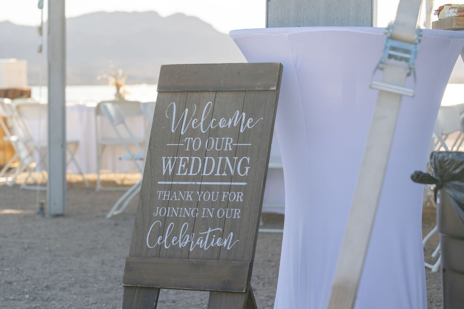 A rustic wooden welcome sign for a wedding celebration stands on a beach with white chairs in the background in Lake Havasu City Arizona