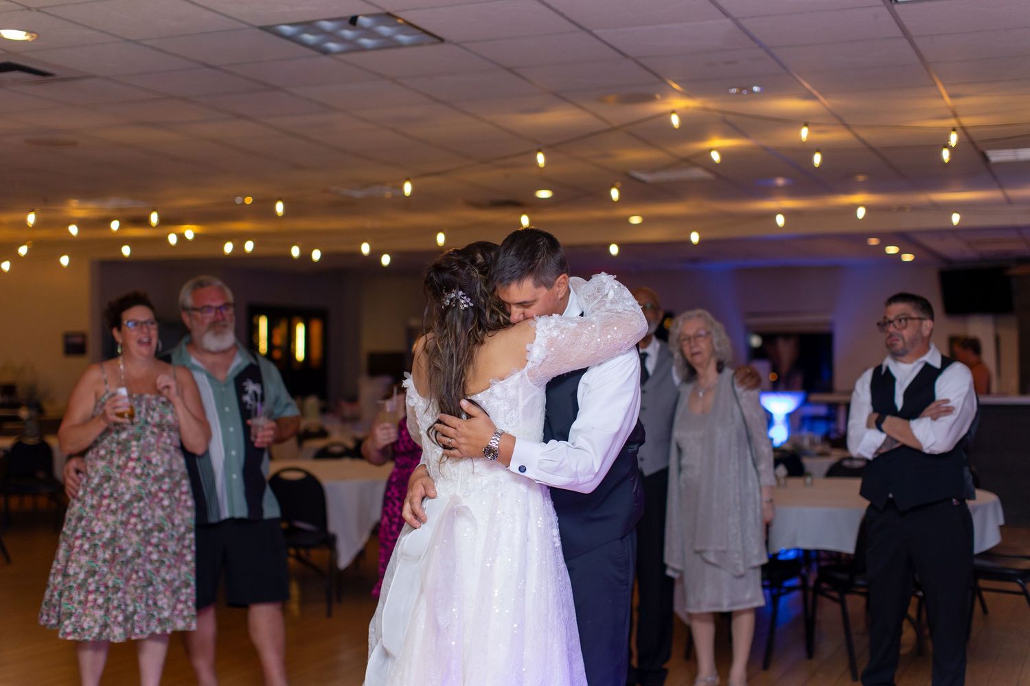 Bride and groom share first dance embrace at wedding reception with string lights and guests watching in background at Shugrues