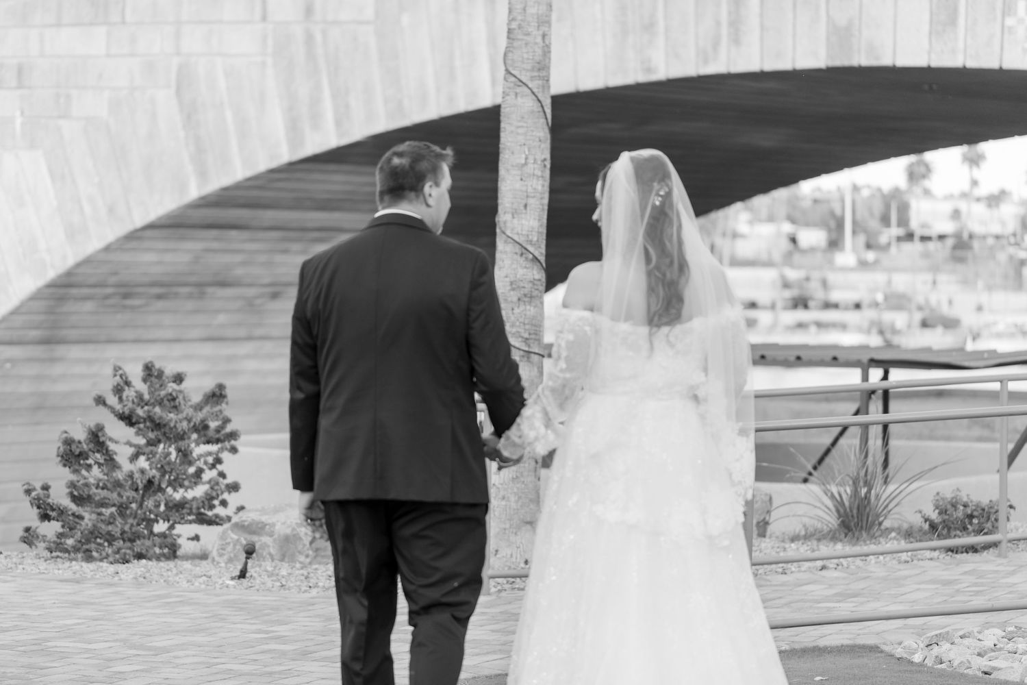 Bride and groom walking hand in hand under an arch in a black and white wedding photo at Shugrues in Lake Havasu by the London Bridge