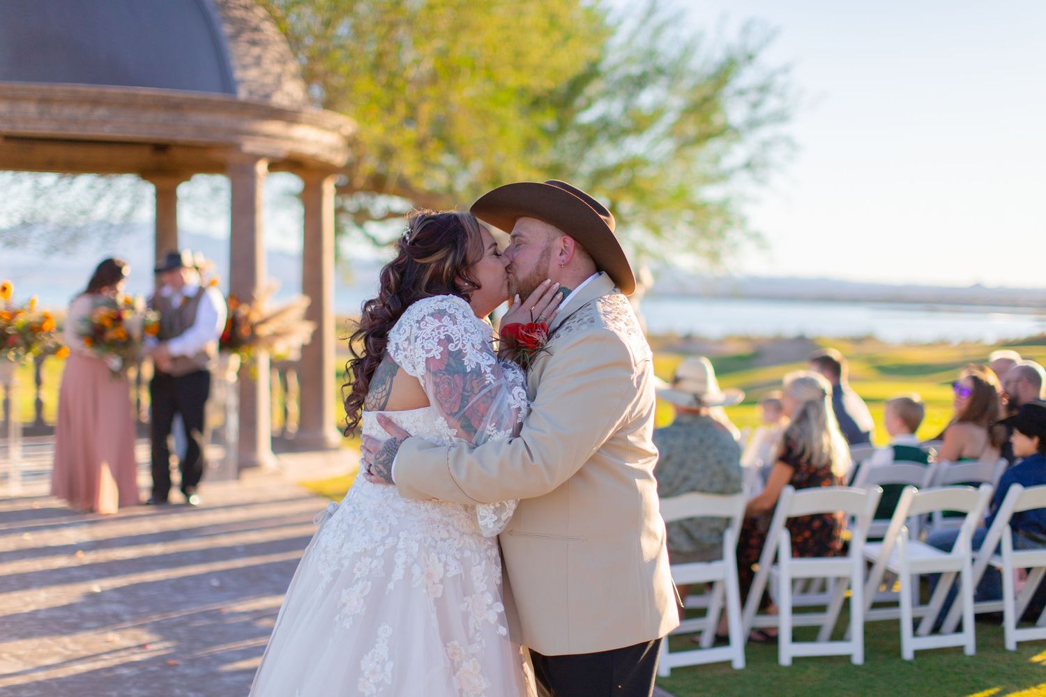 Bride and groom share first kiss at outdoor wedding ceremony near gazebo and lake, groom wearing cowboy hat at Iron Wolf in Lake Havasu