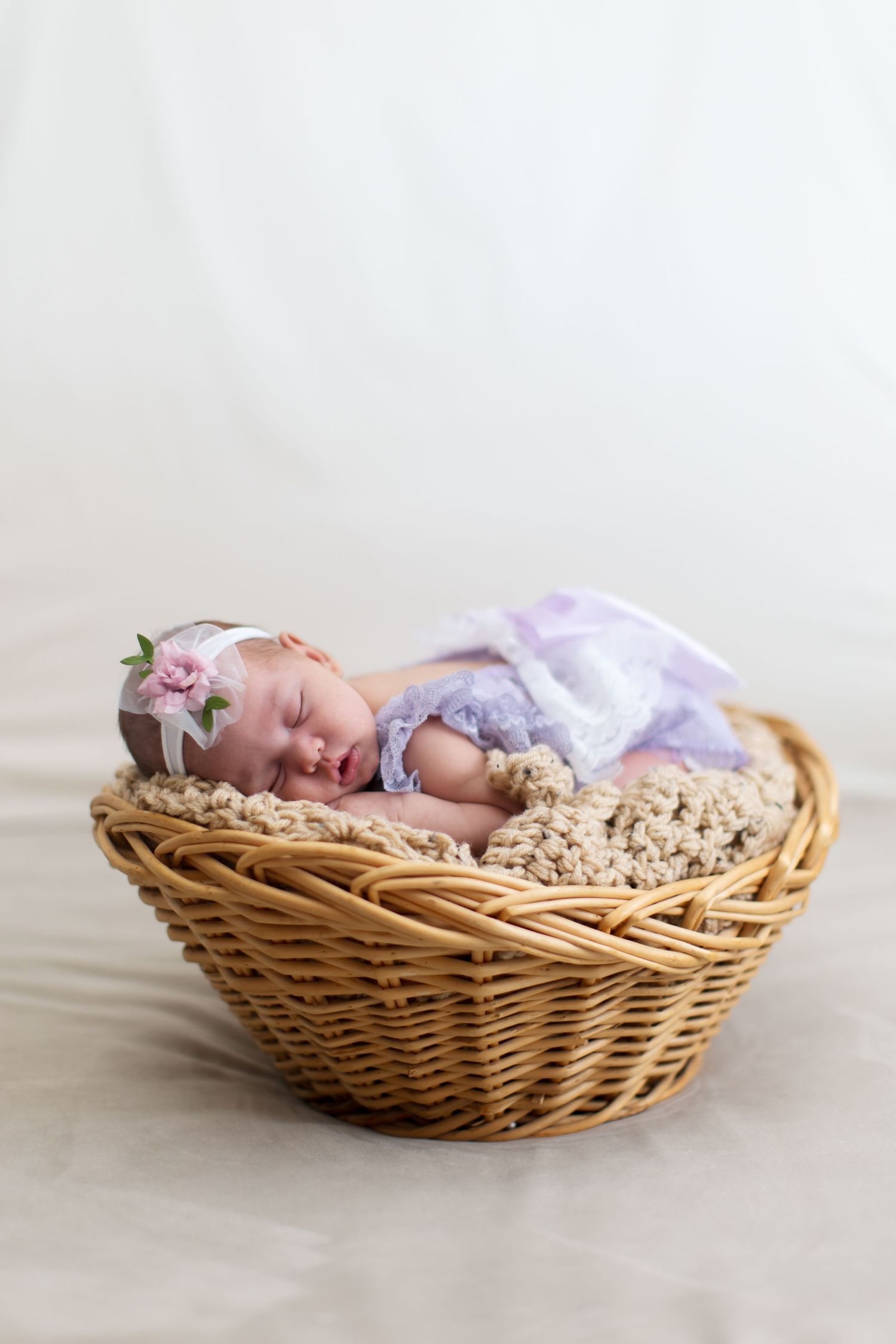 Sleeping newborn baby in purple lace wrap and floral headband nestled in a wicker basket.