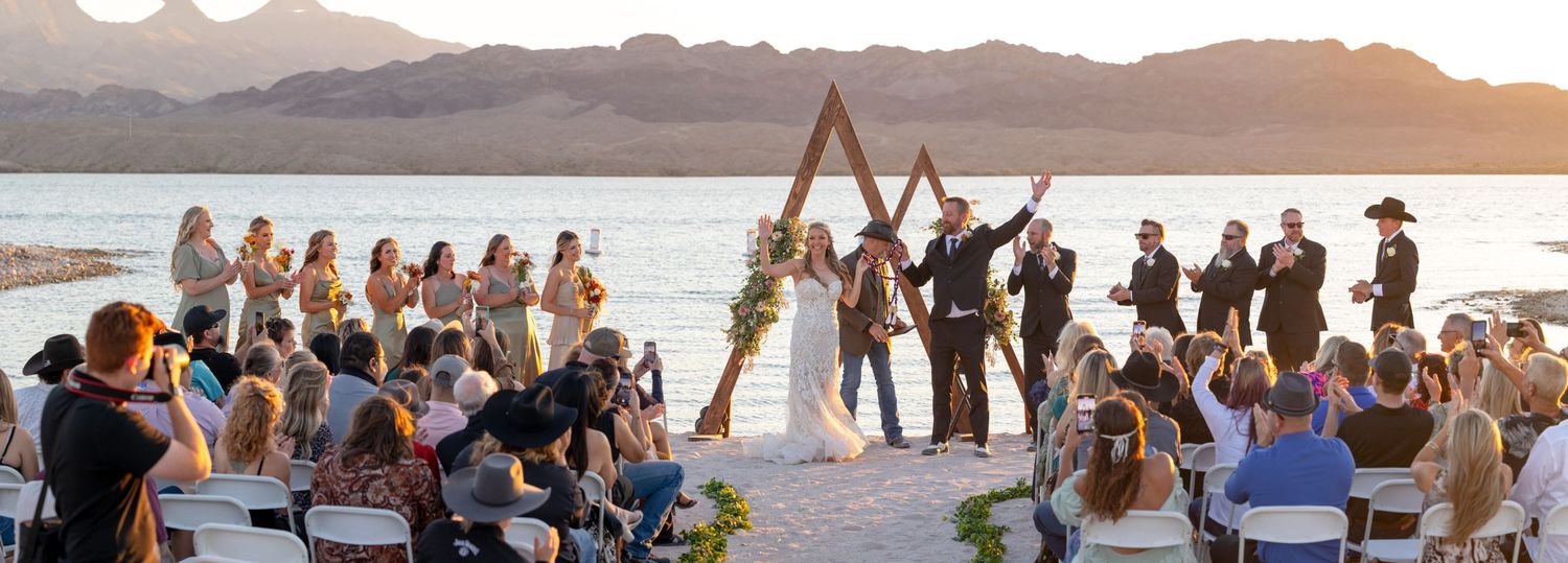 A sunset beach wedding ceremony with guests gathered around a triangular wooden arch overlooking the riviera marina lake havasu