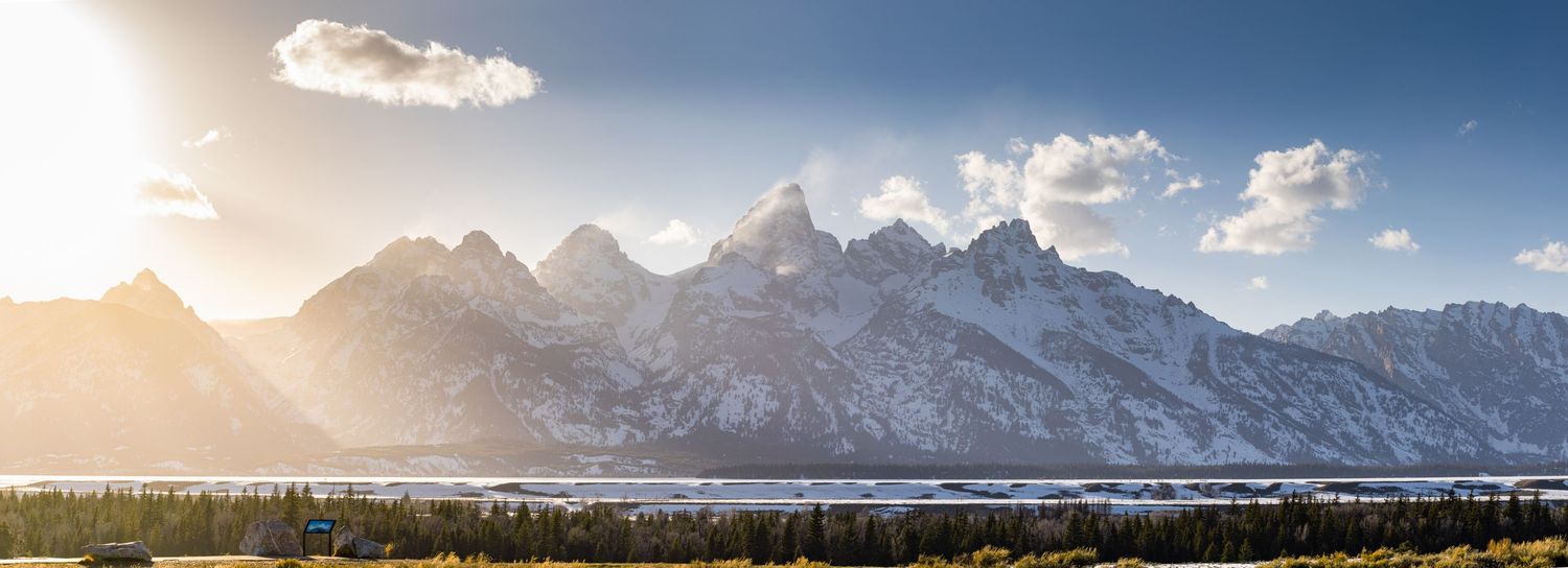 Snow-capped Grand Teton mountain range glowing in golden sunlight with a serene lake and forest in the foreground.