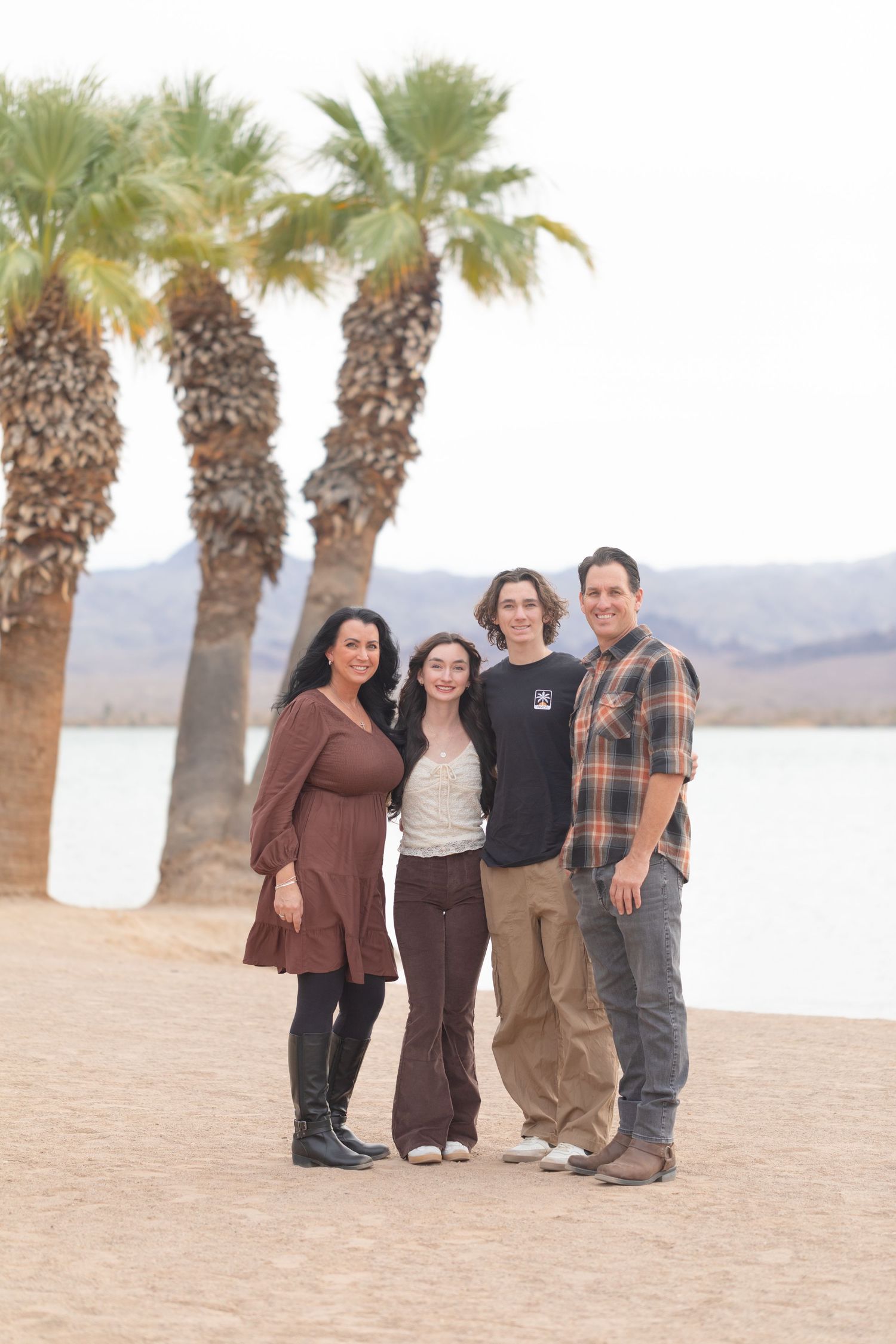 Family of four smiling together on a sandy beach with palm trees and a lake in the background.
