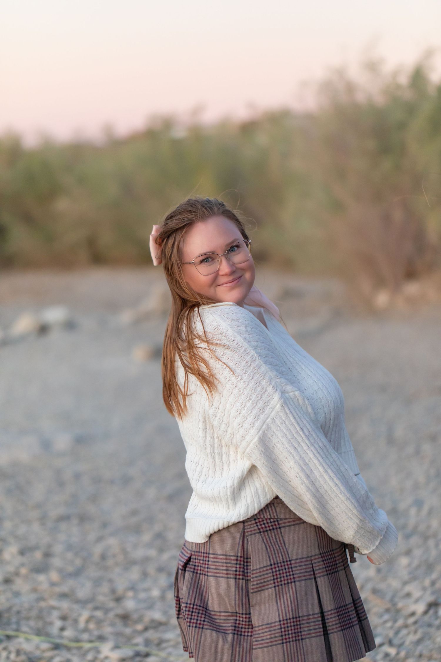 Person in white sweater and plaid skirt poses playfully outdoors.