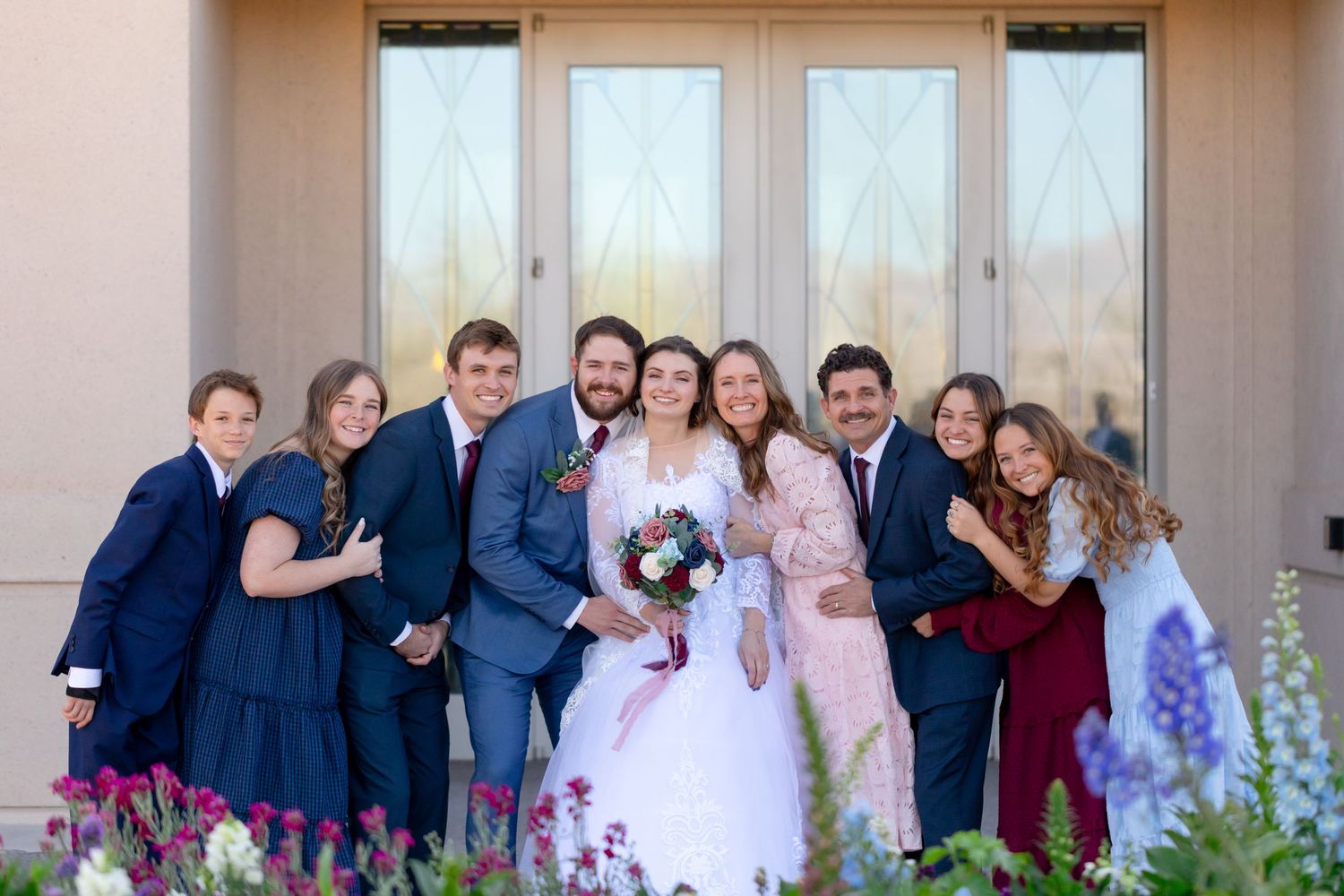 Wedding party poses outside a venue, bride in white gown holding bouquet, surrounded by family in navy and burgundy attire.
