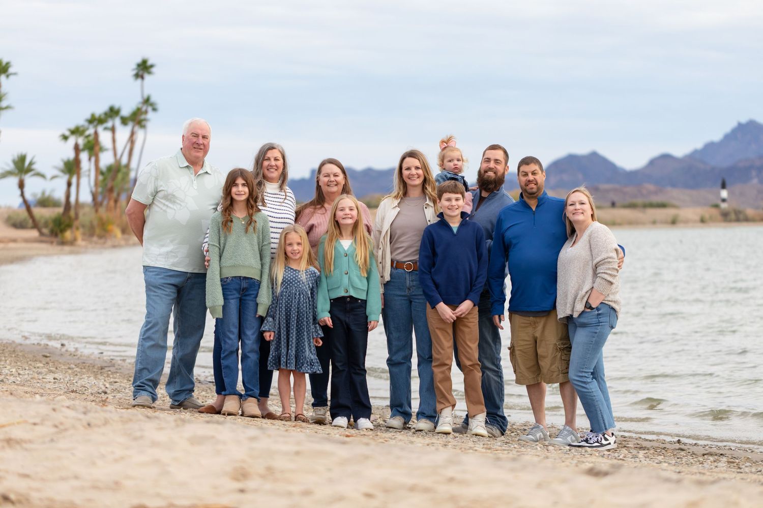 Large family group smiling together on a sandy beach shore with mountains and palm trees in the background.