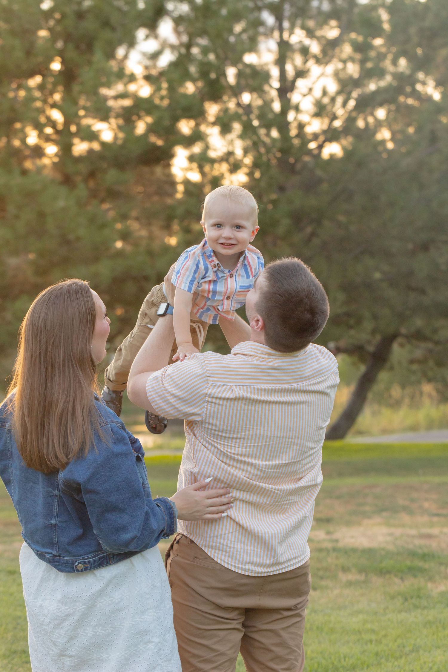 A joyful family moment as parents lift their smiling toddler in a sunny park during golden hour.