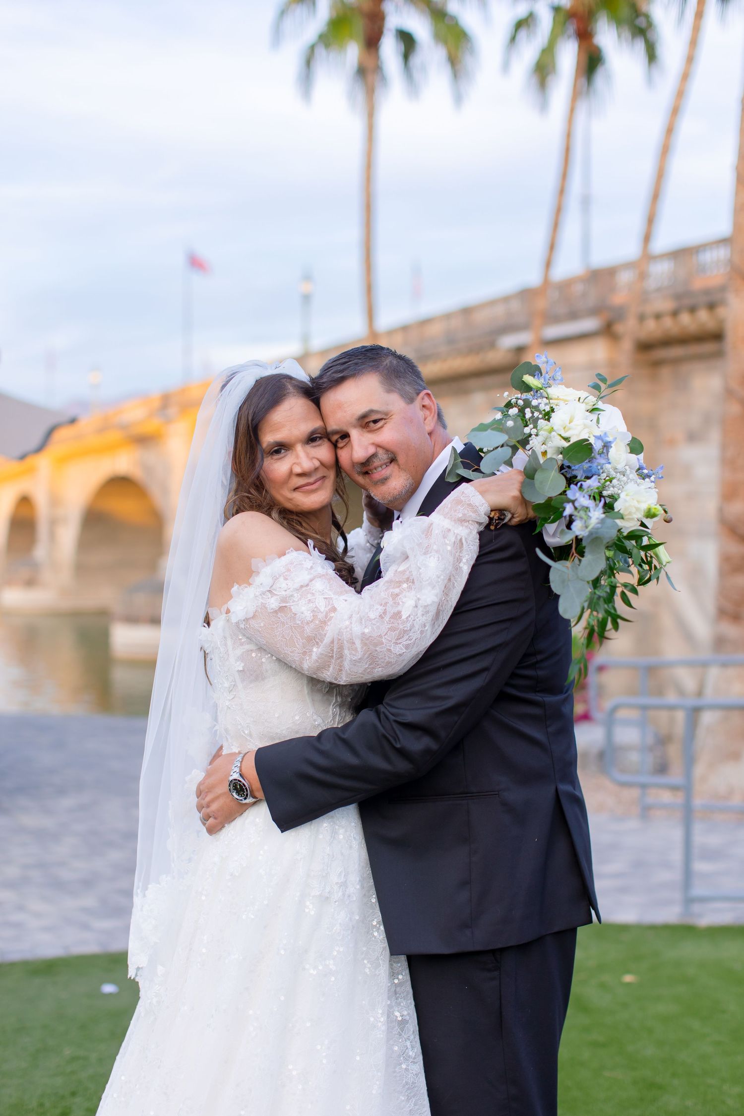 Bride and groom embrace smiling, holding bouquet near palm trees and bridge at waterfront wedding at Shugrues in Lake Havasu by the Bridge