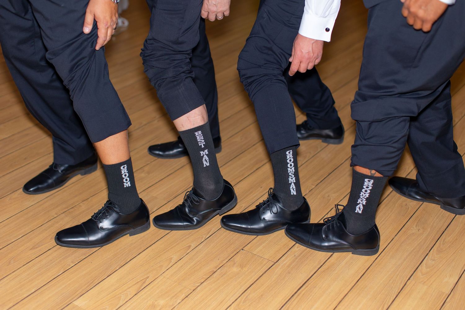 Groomsmen showing off matching custom black dress socks at wedding reception on hardwood floor at Shugrues in Lake Havasu
