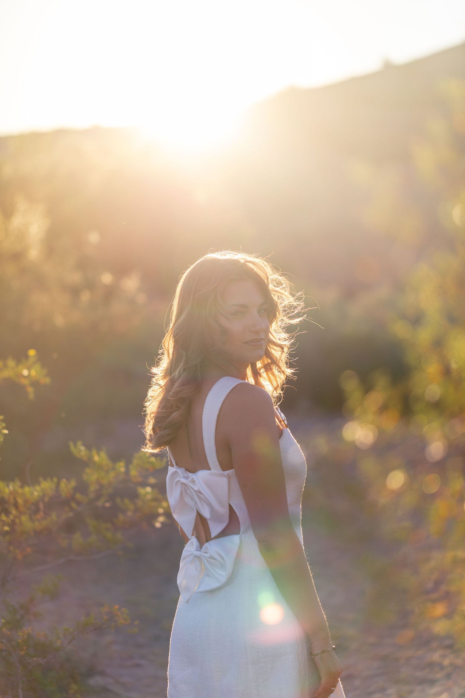 Woman in white backless bow dress glowing in golden sunset light outdoors.