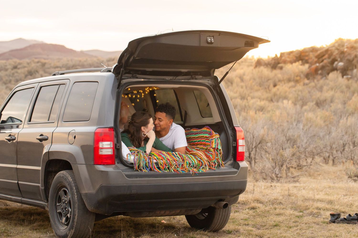 Couple relaxing in the open trunk of a Jeep SUV with string lights, surrounded by scenic mountain landscape at sunset.
