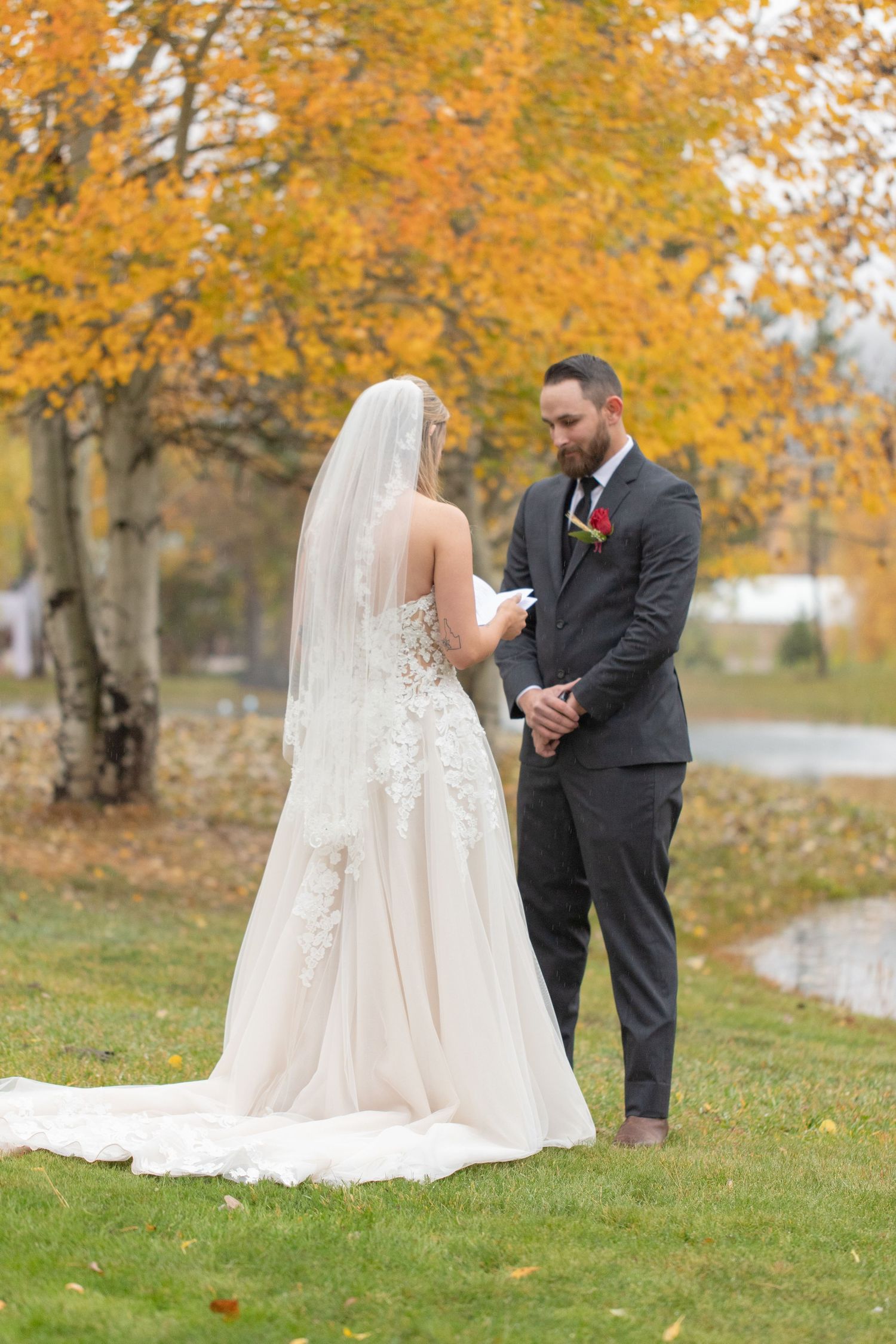 Couple exchanging vows on grassy lawn with fall colors in background.