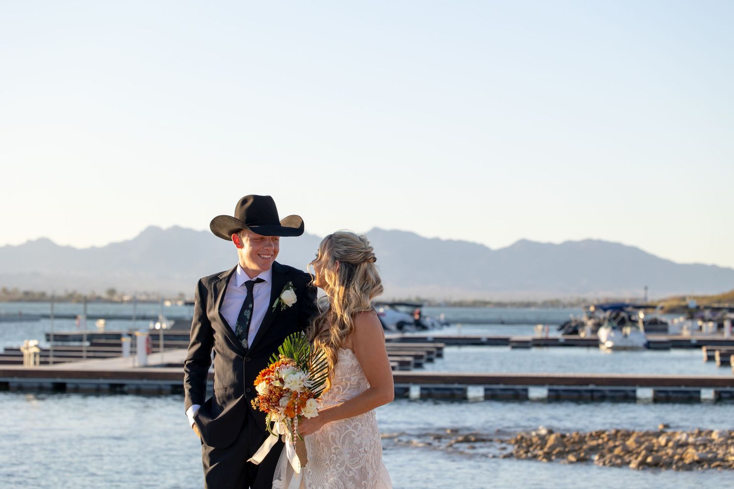 Couple in western attire poses at sunset by Riviera marina with mountains in background and autumn bouquet in Lake Havasu City Arizona