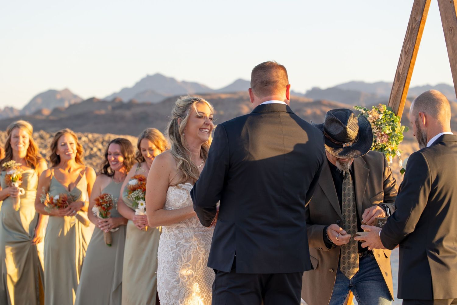 A wedding ceremony takes place outdoors with a scenic mountain backdrop as bridesmaids look on during sunset in Lake Havasu Riviera Marina