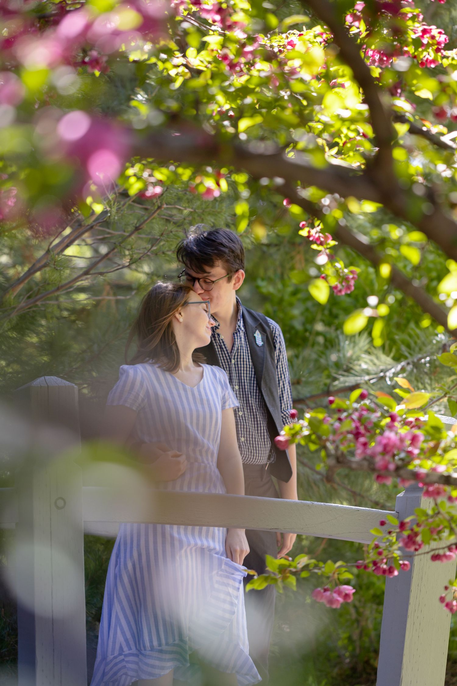 Romantic couple sharing a tender moment on a garden bridge surrounded by blooming pink flowers and lush green trees.