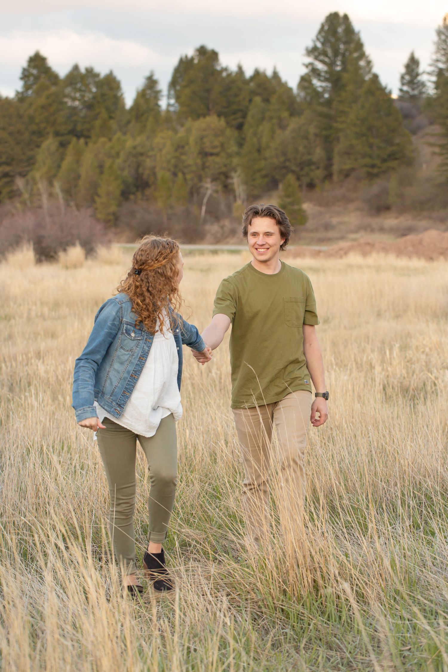Two people walking together through a tall grass meadow with evergreen trees in the background on a sunny day.