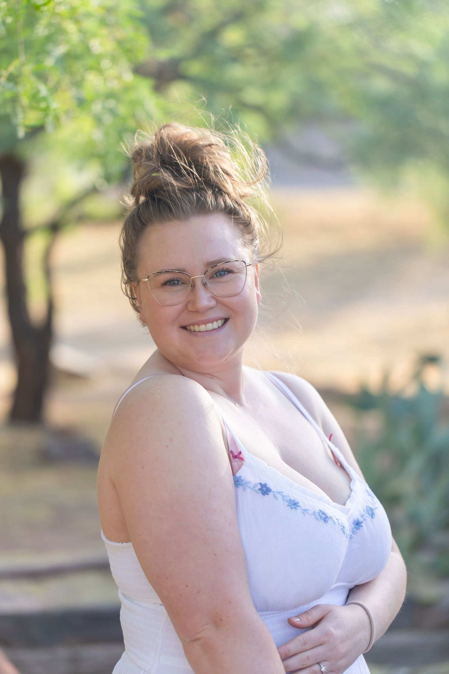 Person in white tank top smiles warmly in outdoor natural setting.