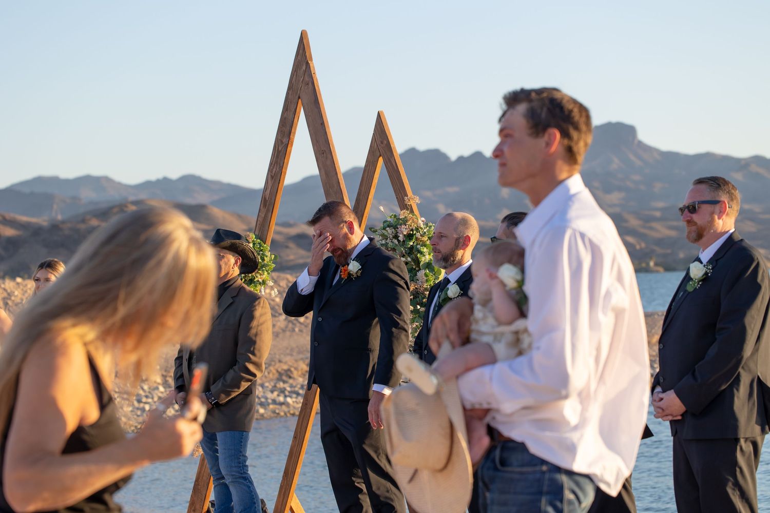 Outdoor wedding ceremony by a lake with mountains, guests watching groom near wooden arch decorated with greenery in Lake Havasu City Arizon