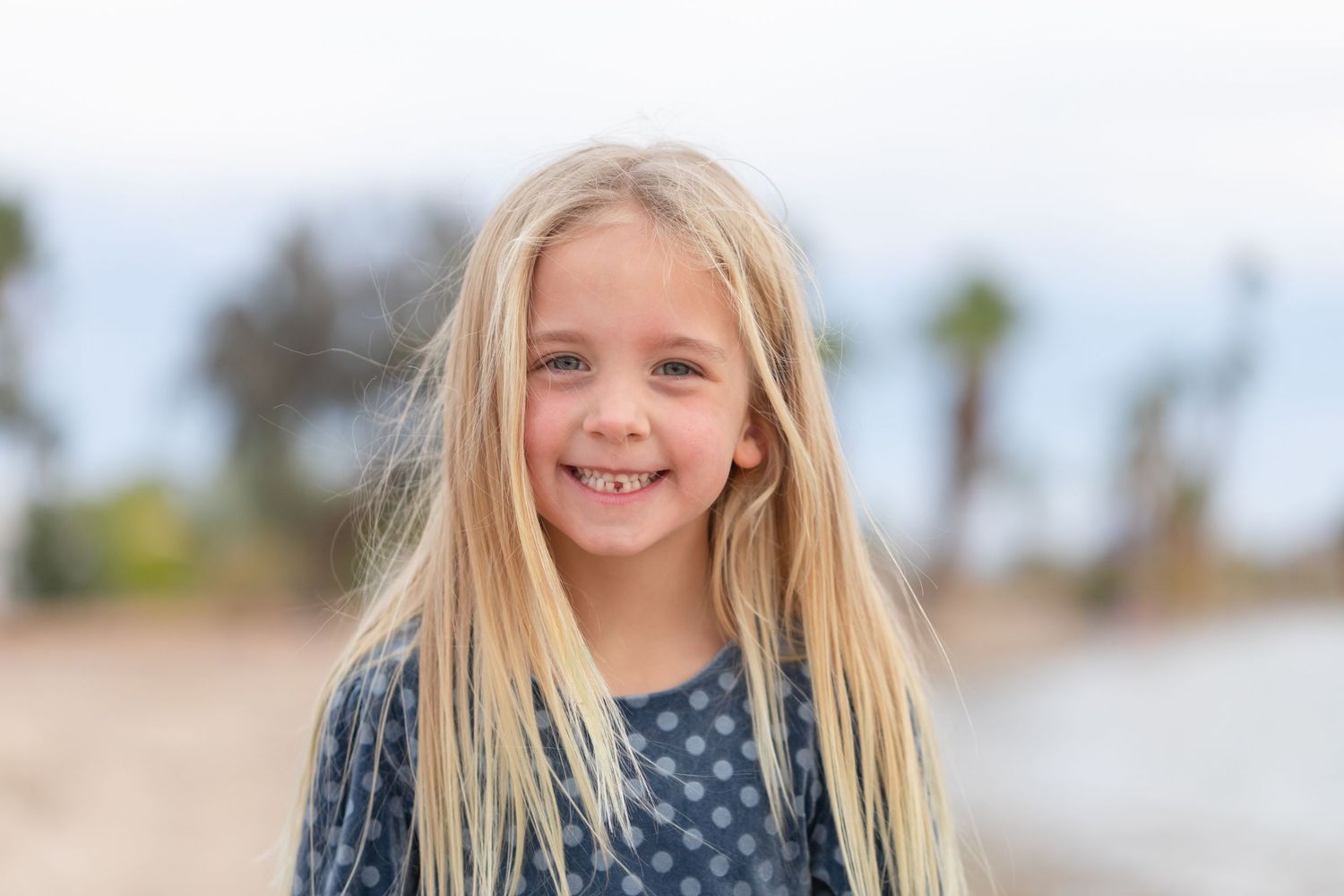 Smiling blonde girl with long hair poses outdoors near a beach with palm trees in the background.
