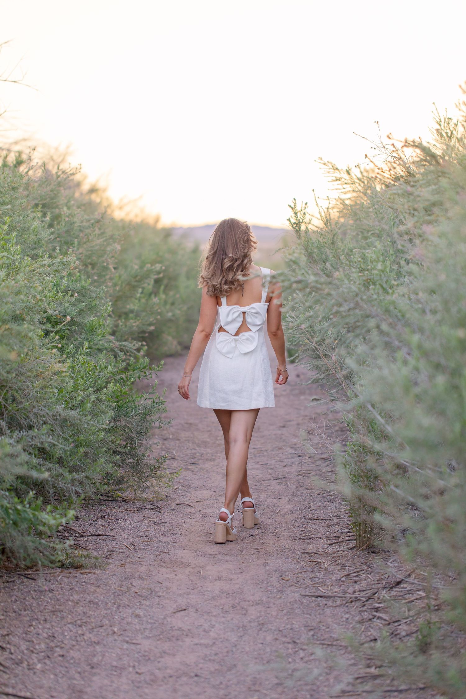 Woman in white bow-back mini dress walking down a scenic dirt path surrounded by lush green shrubs at sunset in Lake Havasu City Arizona