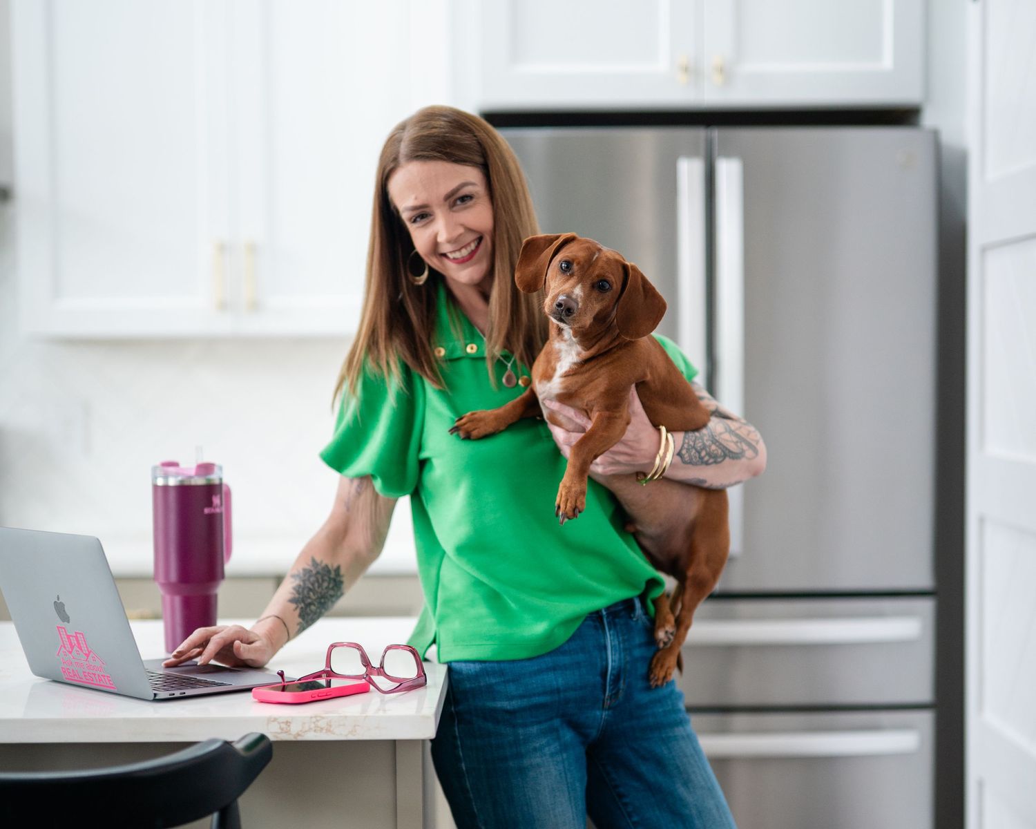 Smiling woman in green scrubs holds a dachshund while working on a laptop in a modern kitchen.