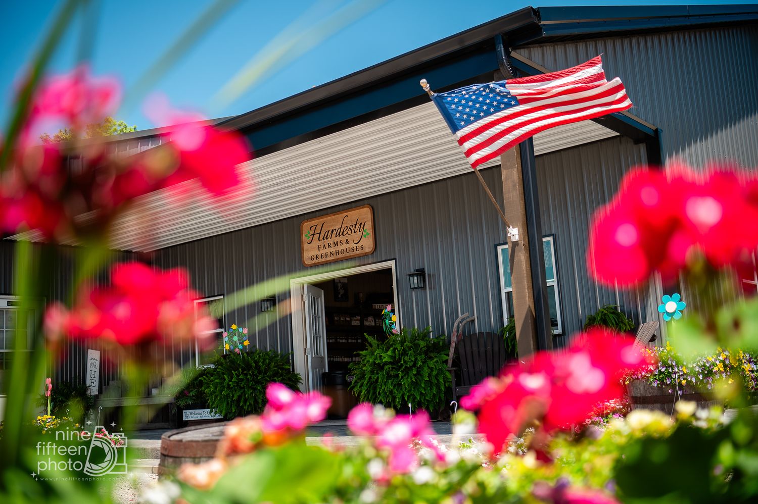 American flag flying above gray farm market building framed by vibrant red flowers at Hardesty Farms.